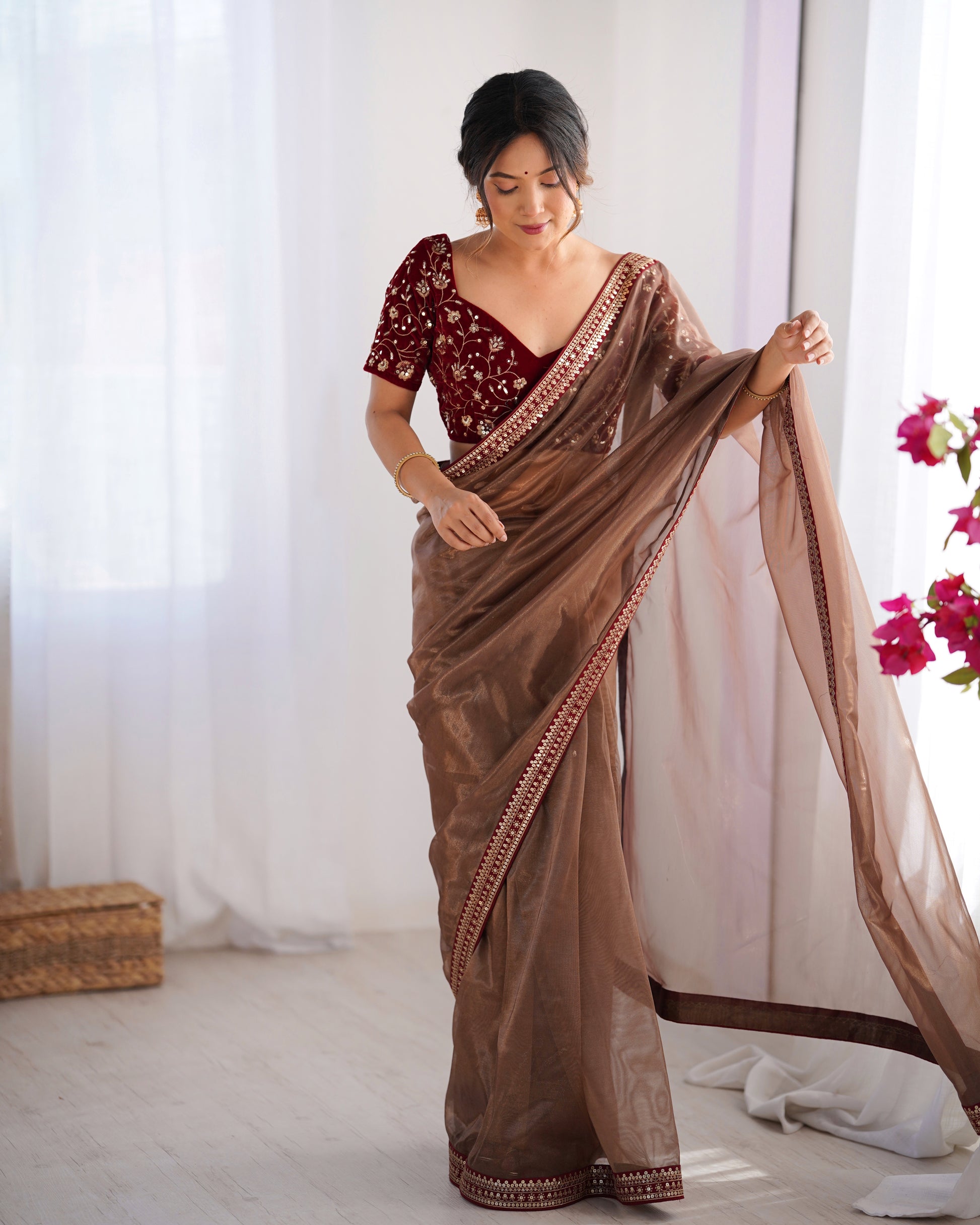 Woman wearing a brown saree with a red blouse, standing indoors with white curtains and flowers in the background.