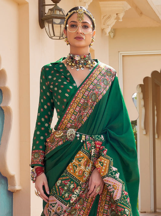 A woman models a traditional Indian green saree with intricate multicolor floral and geometric patterns, paired with a matching embroidered green blouse, ornate jewelry, and a gold headpiece in an architectural corridor.